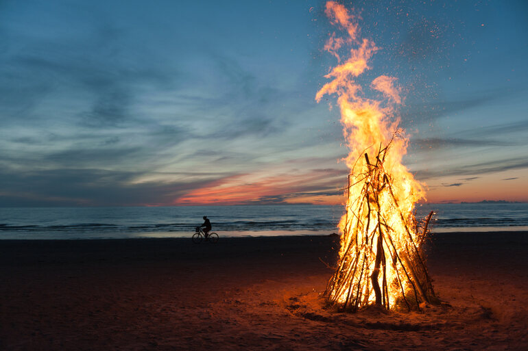 A towering bonfire glows on a beach at dusk while a cyclist silhouetted against the sunset rides by, capturing the desert-festival aesthetic of Burning Man