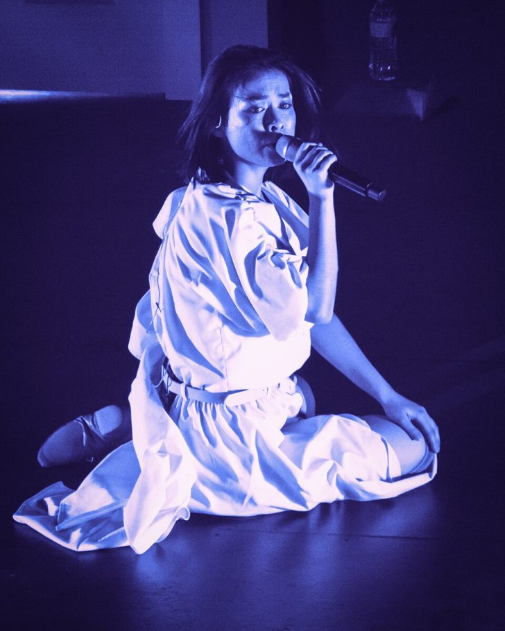 Mitski lying on stage in moody blue lighting with flowing white dress
